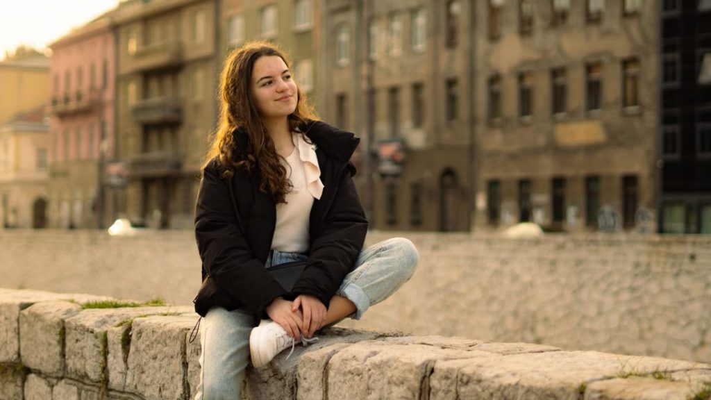 Relaxed young woman with long hair sitting on a stone wall in a cityscape.