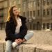 Relaxed young woman with long hair sitting on a stone wall in a cityscape.